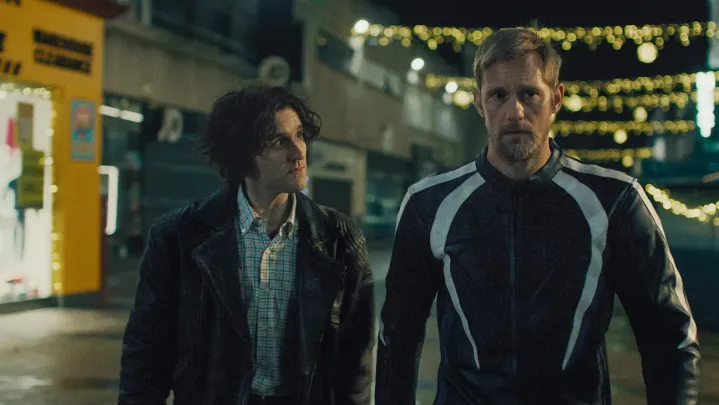 a still from pillion that shows Colin, portrayed by Harry Melling, looking at Ray, portrayed by Alexander Skarsgard